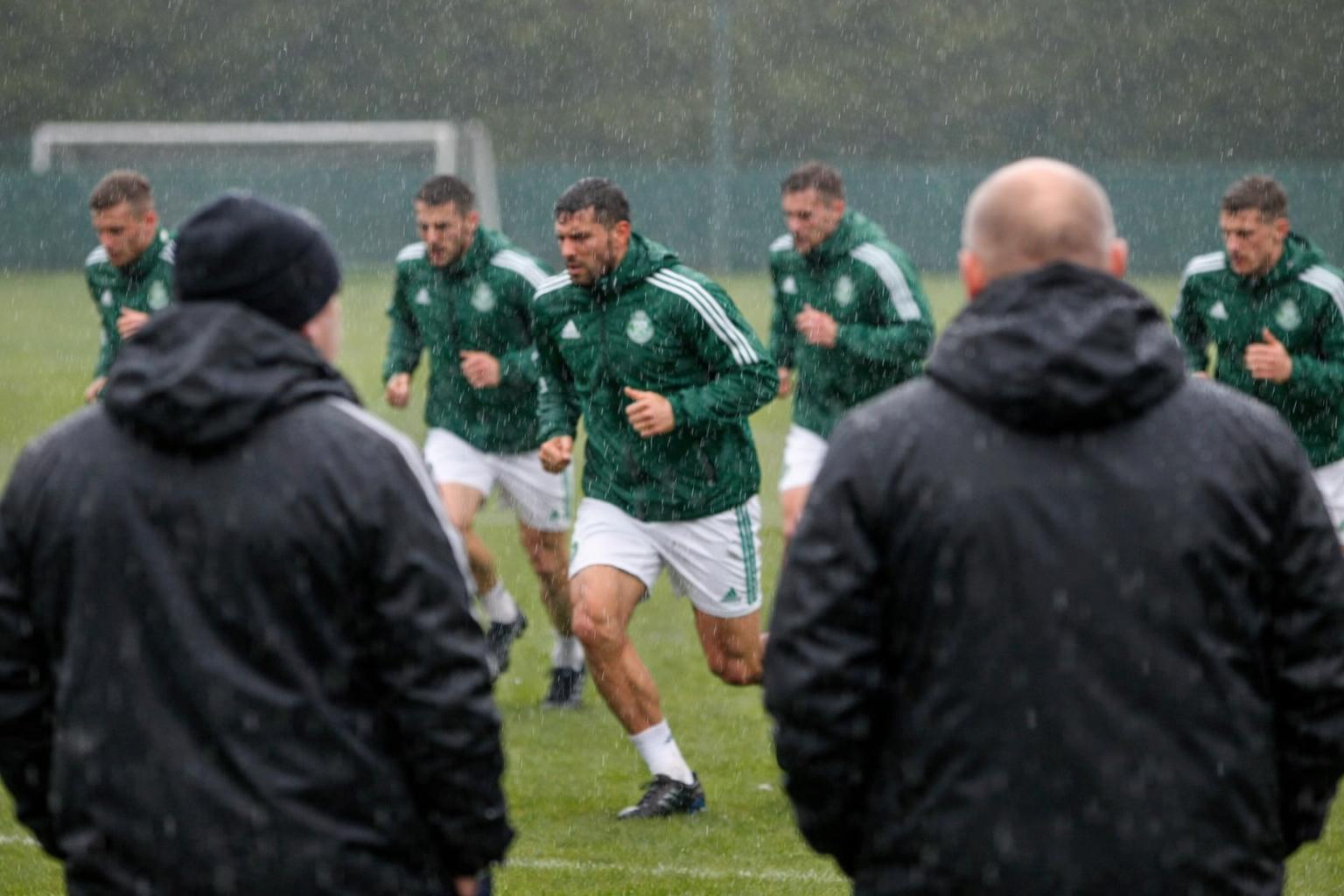 Can I have an image of footballer in Celtic-like tracksuits on the Celtic-like training pitch, running in the rain whilst coa
