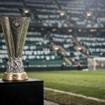 Give me the Europa League trophy sitting on a plinth at the side of the pitch with green and white scarves in the stands