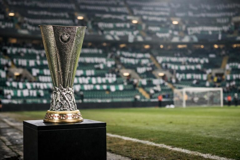 Give me the Europa League trophy sitting on a plinth at the side of the pitch with green and white scarves in the stands